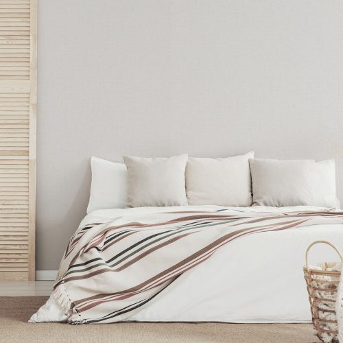 Bedroom with Woven Hessian Wallpaper White on the wall with a side wooden shutter. Large bed draped with blanket and cushions in the foreground.