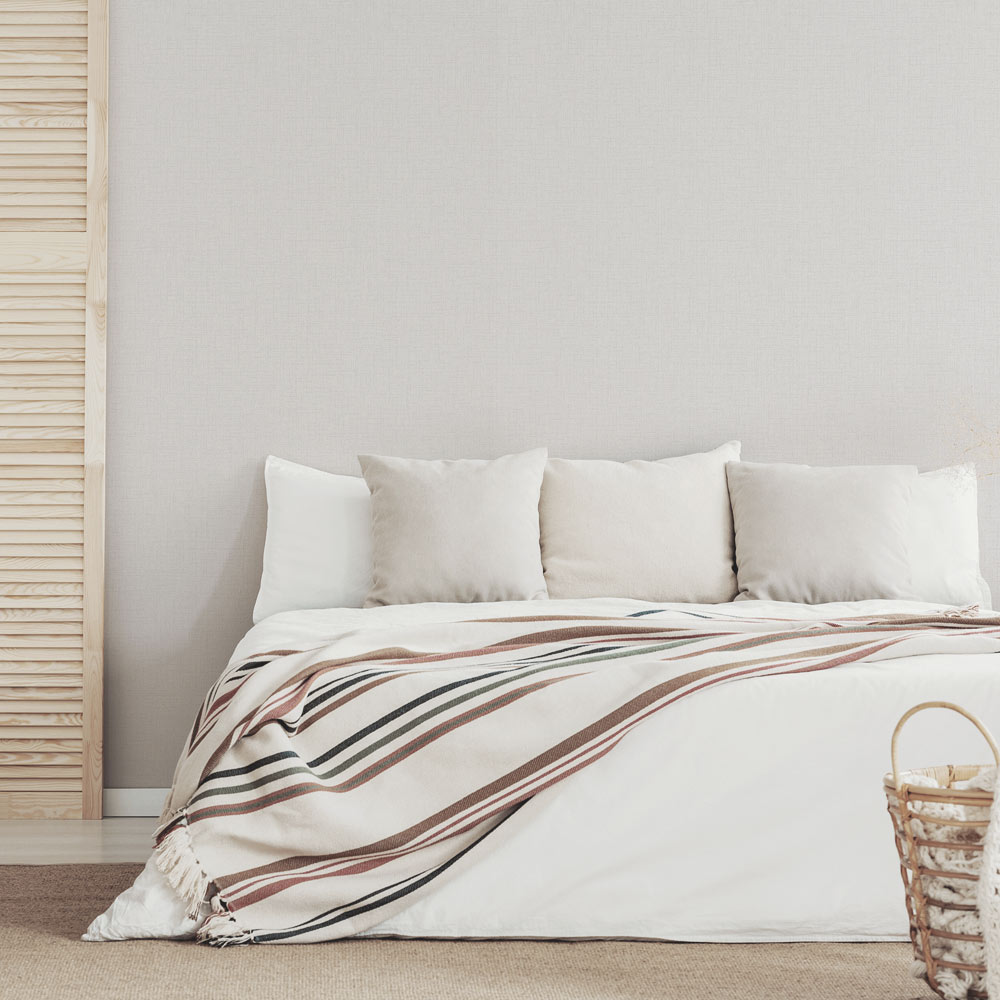 Bedroom with Woven Hessian Wallpaper White on the wall with a side wooden shutter. Large bed draped with blanket and cushions in the foreground.