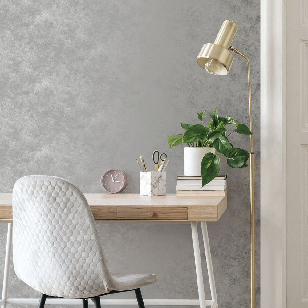 Home office with the metallic concrete wallpaper pearl installed on the wall. Also features a light wood desk with plant, pen holder, books and desk clock - as well as silver velvet desk chair and gold standing lamp