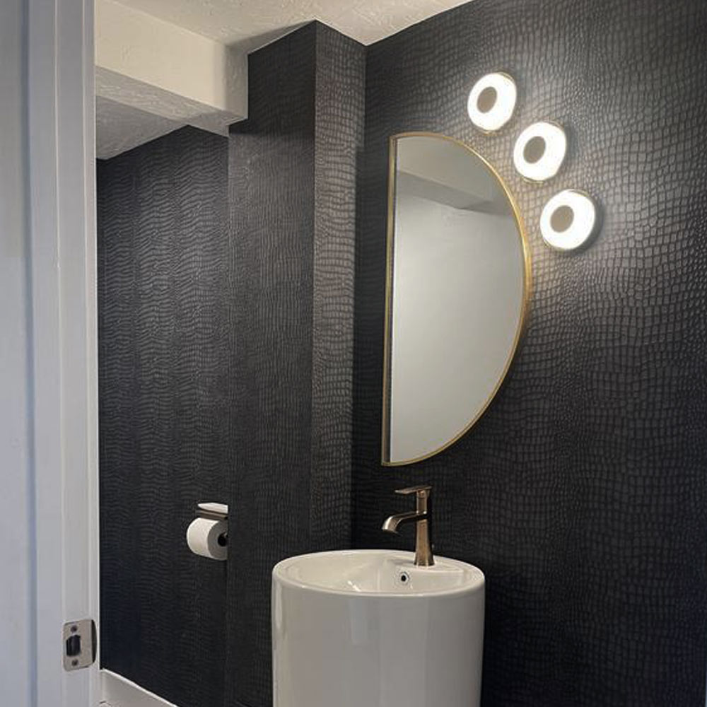 Bathroom with Crocodile Wallpaper - Black installed onto the walls. Bathroom has a white pedestal basin, half moon mirror and 3 wall lights accentuating the wallpaper pattern.