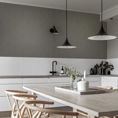 Kitchen with Metallic Draped Silk Wallpaper Smokey Quartz installed on the wall above white tiles. Kitchen also features black pendant lamps, black tap with sink and wooden table and chairs