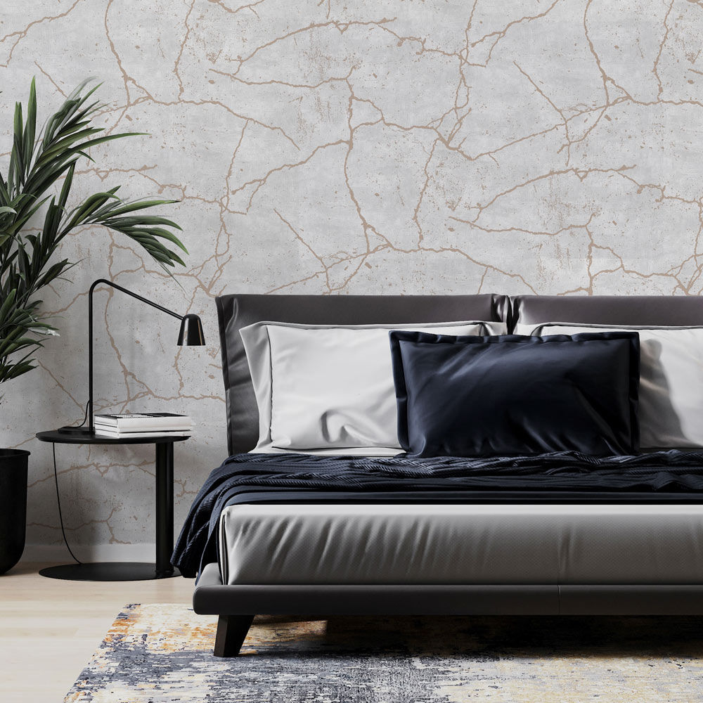 Bedroom with textured concrete cracks metallic wallpaper on the wall. Also features a large grey bed with pillows, side table with desk lamp plus potted palm