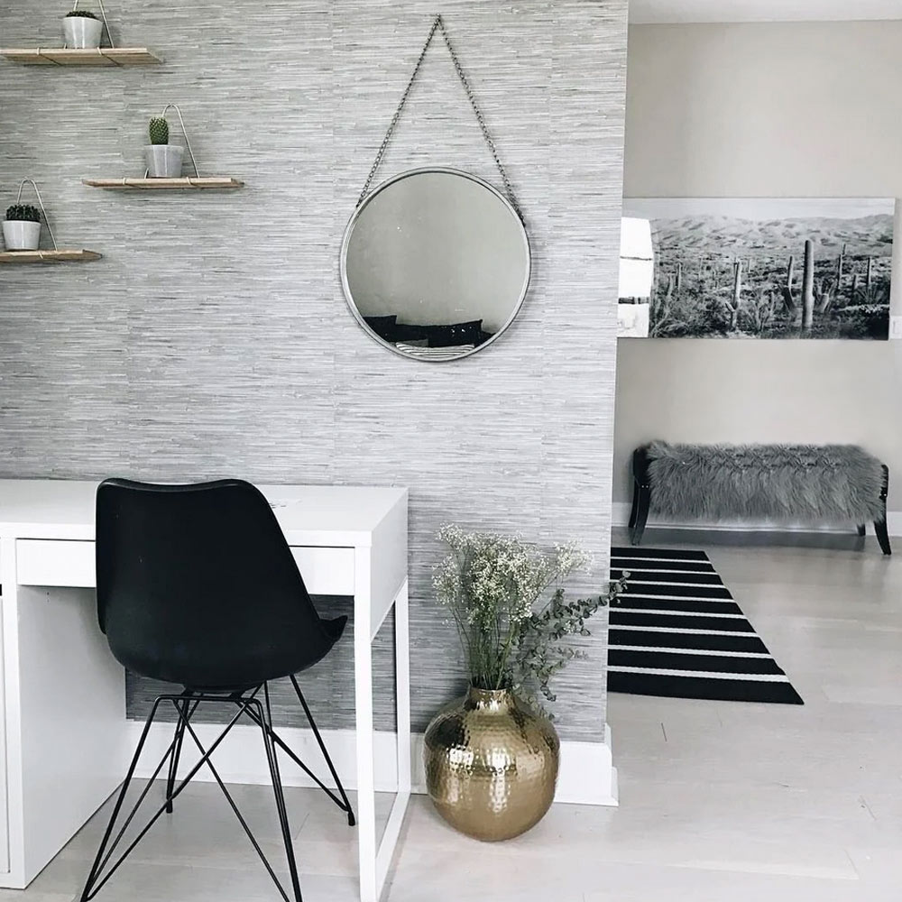 Home office with woven grasscloth wallpaper grey installed on the wall. Also features a small desk, black chair, wall mirror and shelves. Background shows a couch, mat and monochrome picture on wall.