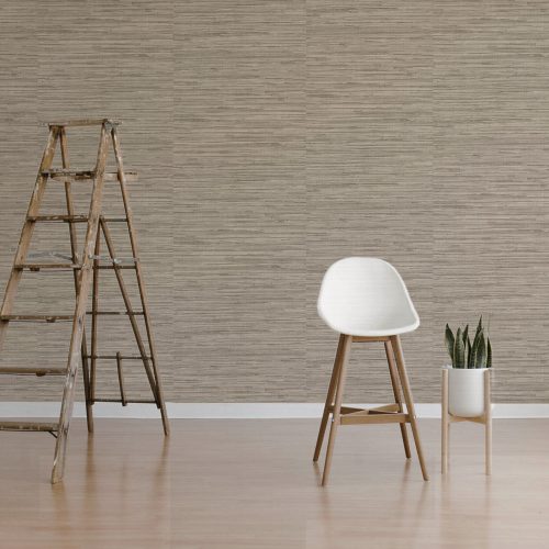 Styled room shot of the woven grasscloth wallpaper warm brown installed on the wall. The foreground fetaures a high scandanavian chair, plant in stand and ladder